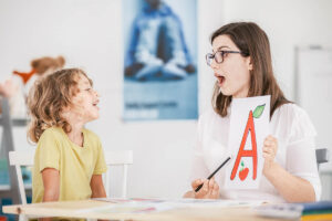 Speech therapist working with a child on a correct pronunciation using a prop with a letter 'a' picture.
