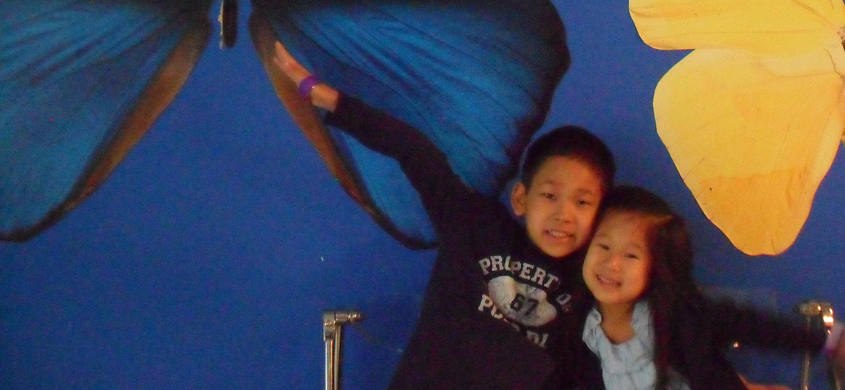 two young children siblings with their arms around each other in front of a wall with a large blue and yellow butterfly