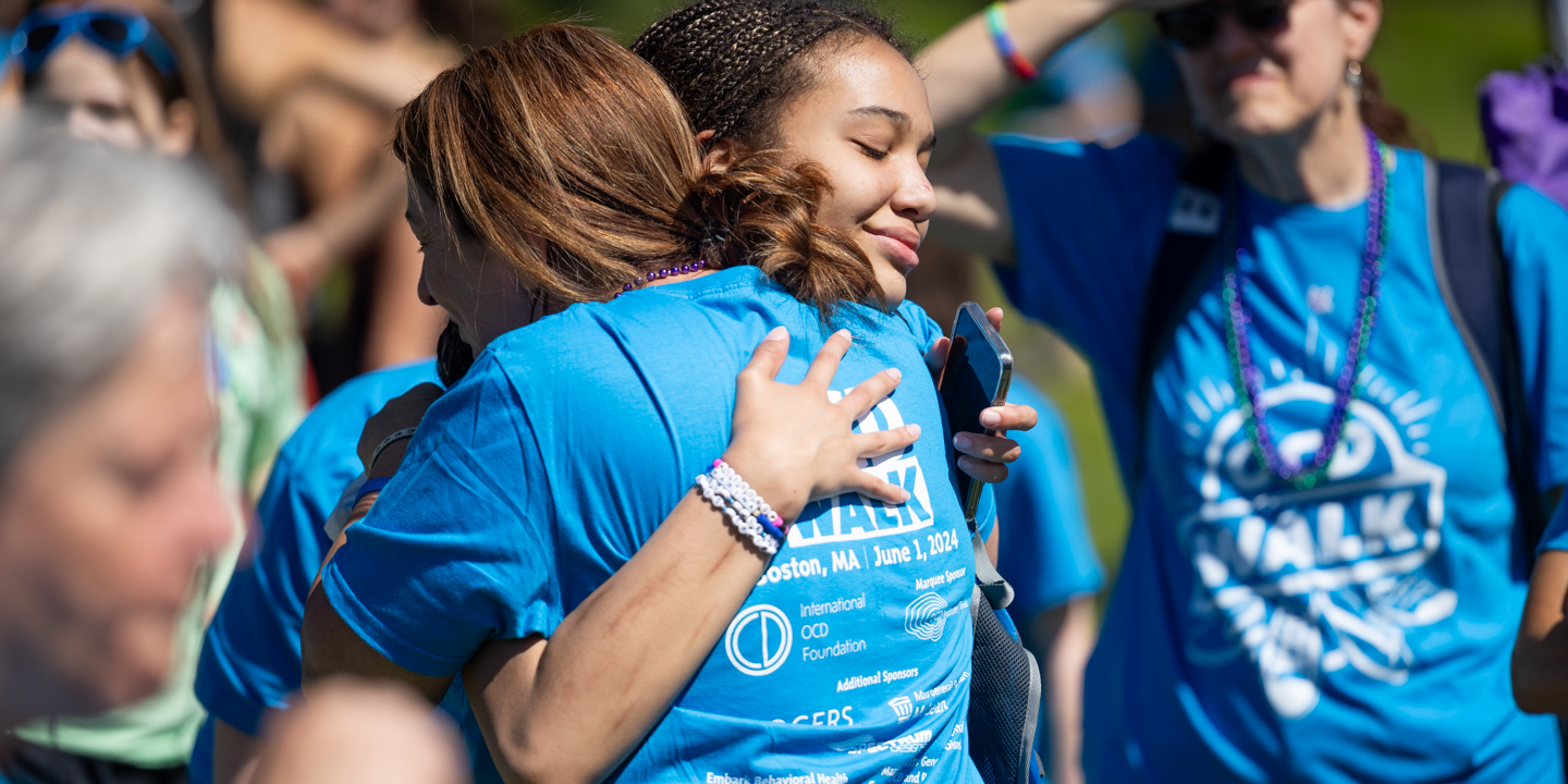 Two young women wearing cyan OCD Walk t-shirts embracing at the Boston OCD Walk in a crowd of people